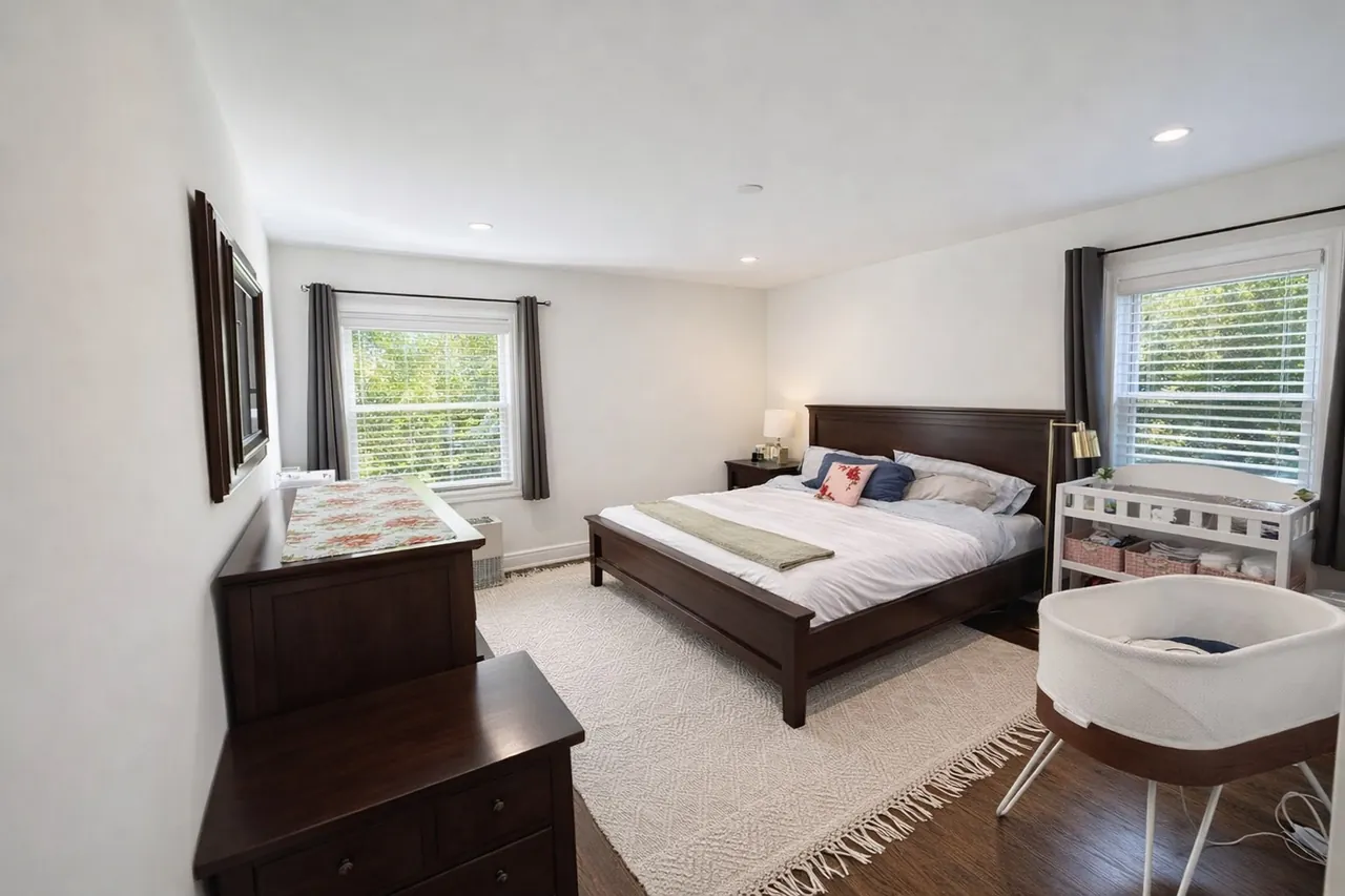 Warm, bright master bedroom with dark walnut furniture, a bassinet and crib, and two daylit windows after photo enhancement