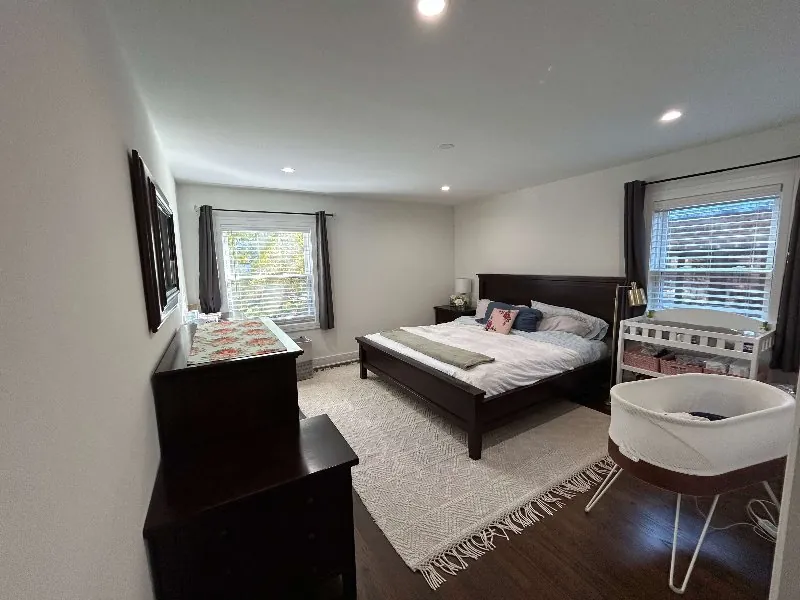 Warm, bright master bedroom with dark walnut furniture, a bassinet and crib, and two daylit windows after photo enhancement (original, before AI enhancement)